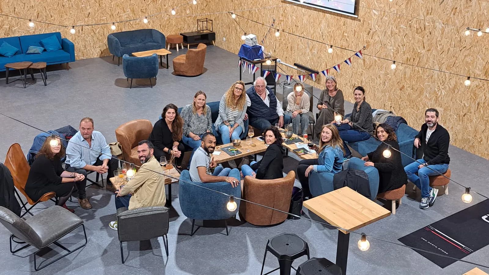 Groupe d'une quinzaine de personnes réunies au Hache Bar pour une soirée d'inauguration : canapés bleus, fauteuils en cuir, guirlandes lumineuses, écran de projection, ambiance team building.